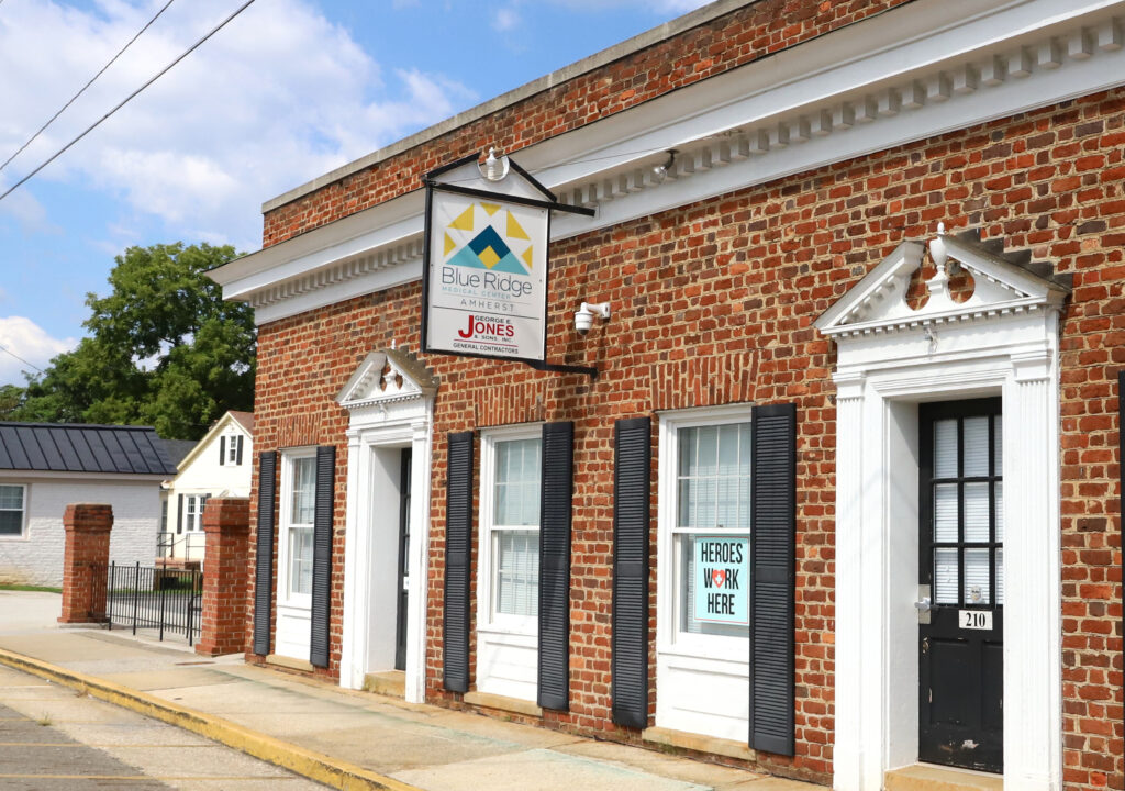 A brick building with white trim houses the Blue Ridge Medical Center sign above the entrance. With black shutters on the windows, one has a sign reading "Heroes Work Here." A sidewalk runs along the front, leading through downtown Amherst, under a partly cloudy sky. - Blue Ridge Medical Center