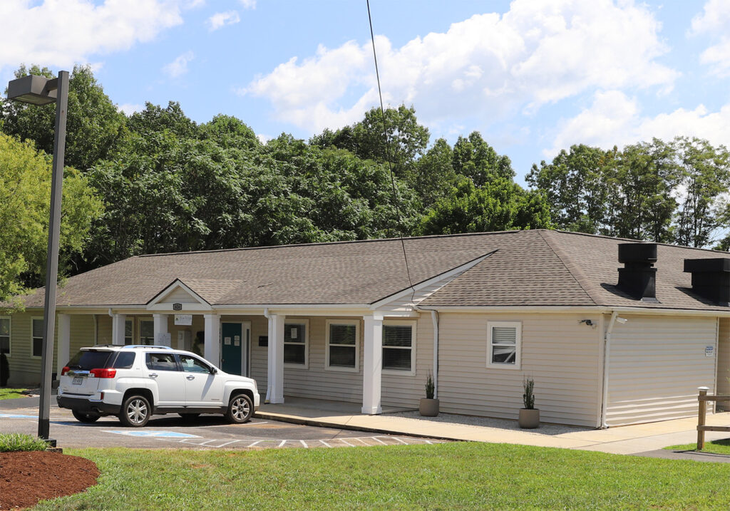 A one-story beige building with white trim, featuring a front entrance with columns. A white SUV, bearing an Appomattox commemorative plate, is parked in front near a few potted plants by the entrance. The area boasts a grassy lawn, and the sky is partly cloudy with trees in the background. - Blue Ridge Medical Center