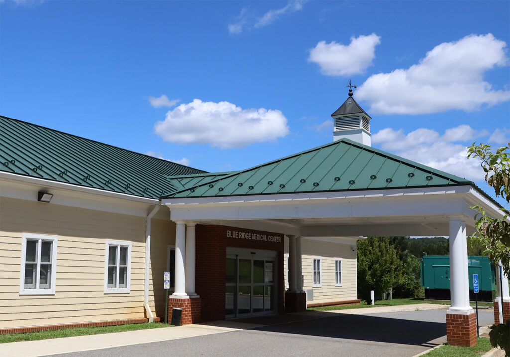 Arington's medical center boasts a green metal roof and beige siding. The entrance features a covered drop-off area supported by white columns and brick bases. A cupola with a weathervane adorns the roof. The sky is blue with scattered clouds, and there are trees in the background. - Blue Ridge Medical Center