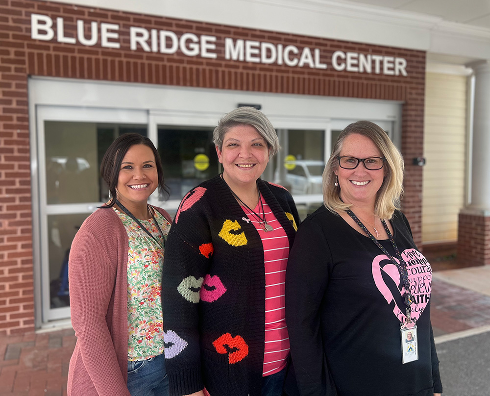 Three smiling women stand in front of the Blue Ridge Medical Center entrance. They are wearing casual clothing, including cardigans and tops with colorful patterns. - Blue Ridge Medical Center