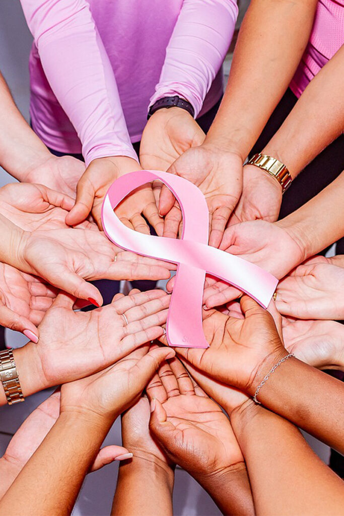 A group of diverse hands joins together, palm up, holding a pink ribbon symbolizing breast cancer awareness. The background is neutral, highlighting the unity and support for the cause. - Blue Ridge Medical Center