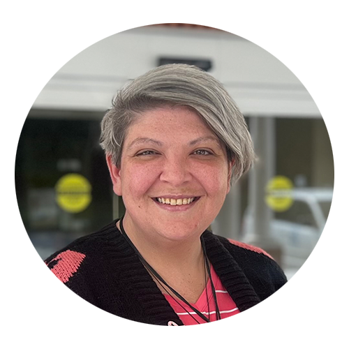 A person with short, gray hair smiles while wearing a black sweater with pink accents over a pink top. They are standing in front of a building with glass doors. - Blue Ridge Medical Center